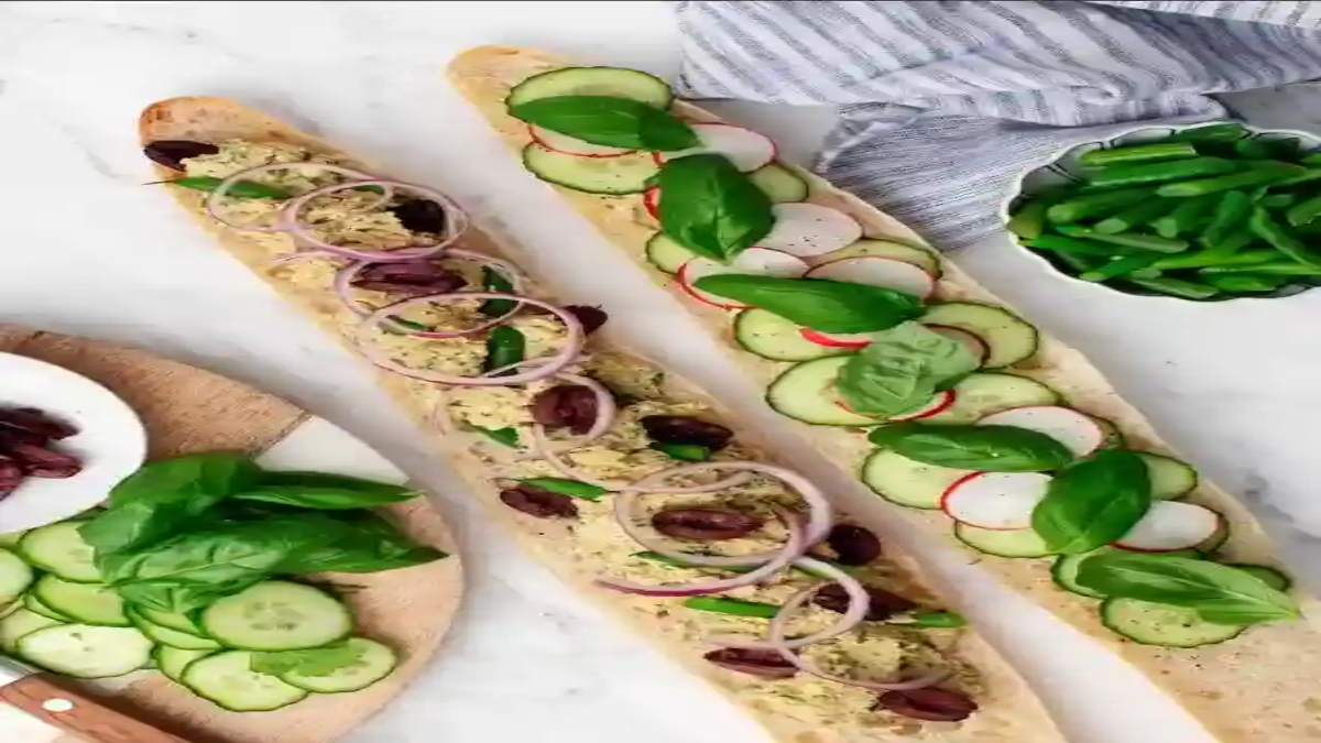 Two open-faced baguette sandwiches topped with sliced cucumbers, radishes, basil, hummus, olives, and red onion on a white surface.