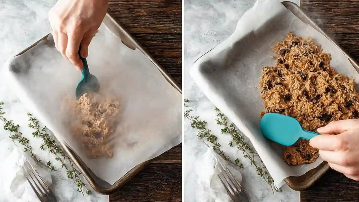 Spreading homemade granola bar mixture with chocolate chips onto a parchment-lined baking tray using a spatula.