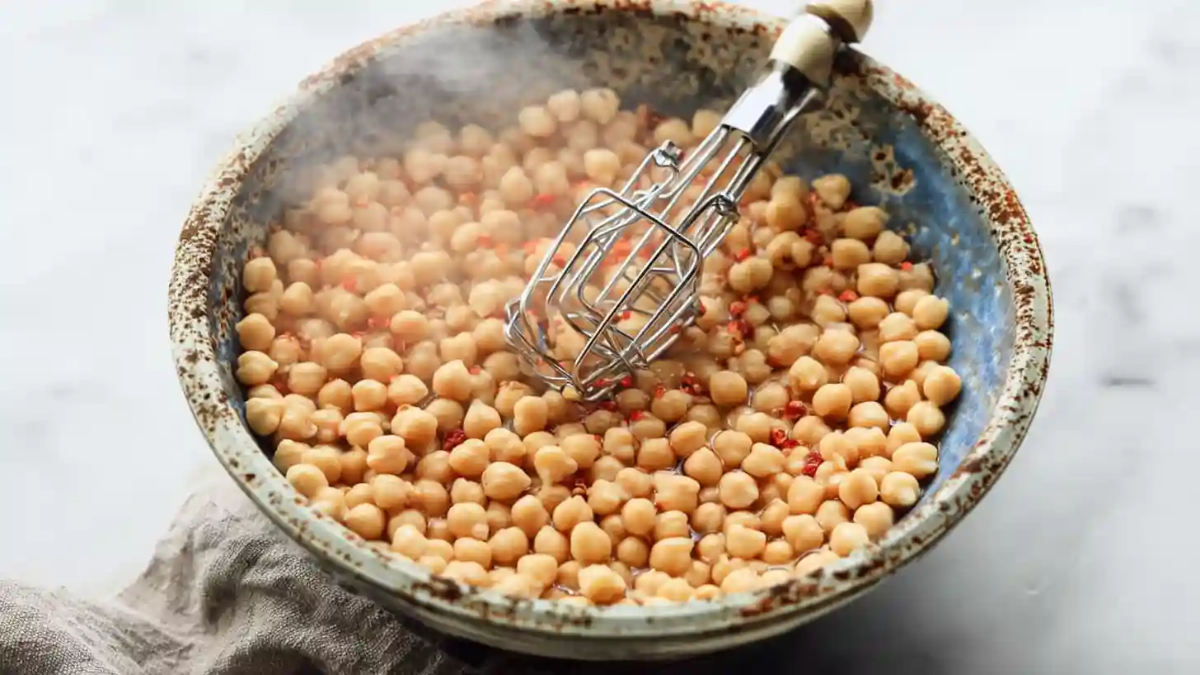 Warm chickpeas with diced red peppers in a rustic bowl being mashed with a handheld masher on a white kitchen surface.