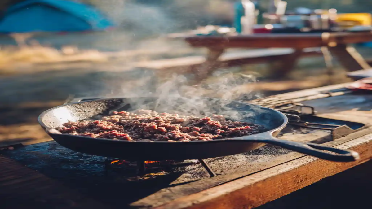 Ground beef sizzling in a cast iron skillet over an open flame at a rustic campsite cooking station.