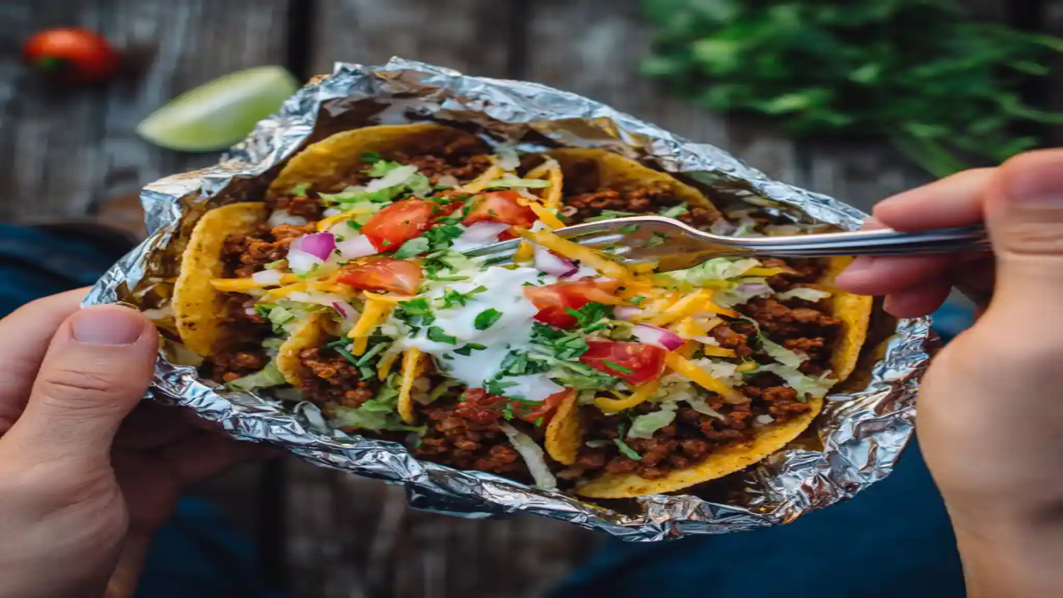 Close-up of foil-wrapped beef tacos with cheese, lettuce, tomato, and sour cream, being eaten outdoors.